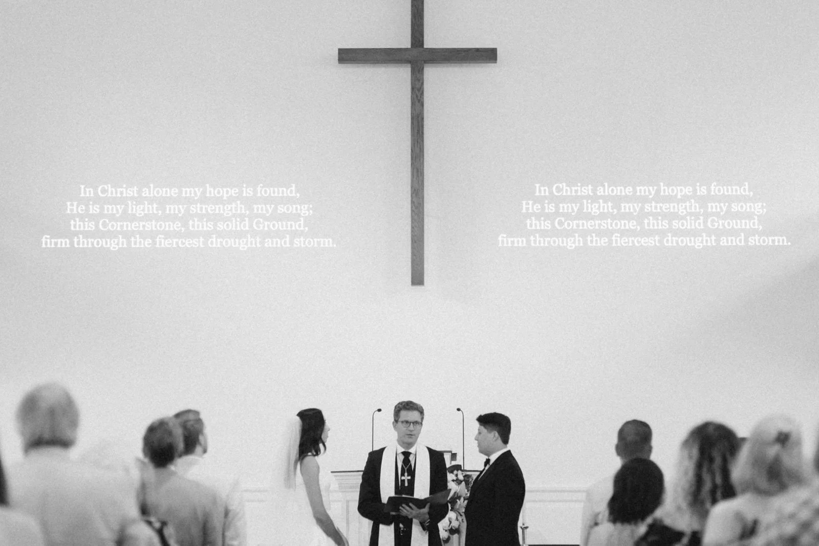 A couple getting married at a church ceremony, with a minister officiating, in front of a cross and religious text on the wall.