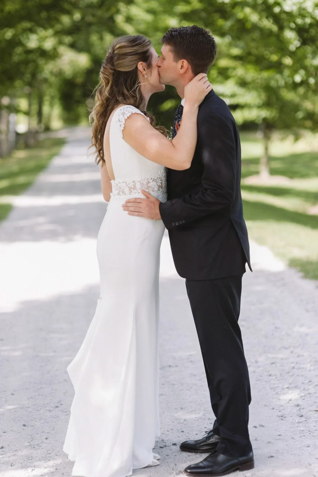 A bride and groom sharing a kiss outdoors on a tree-lined path, dressed in wedding attire.