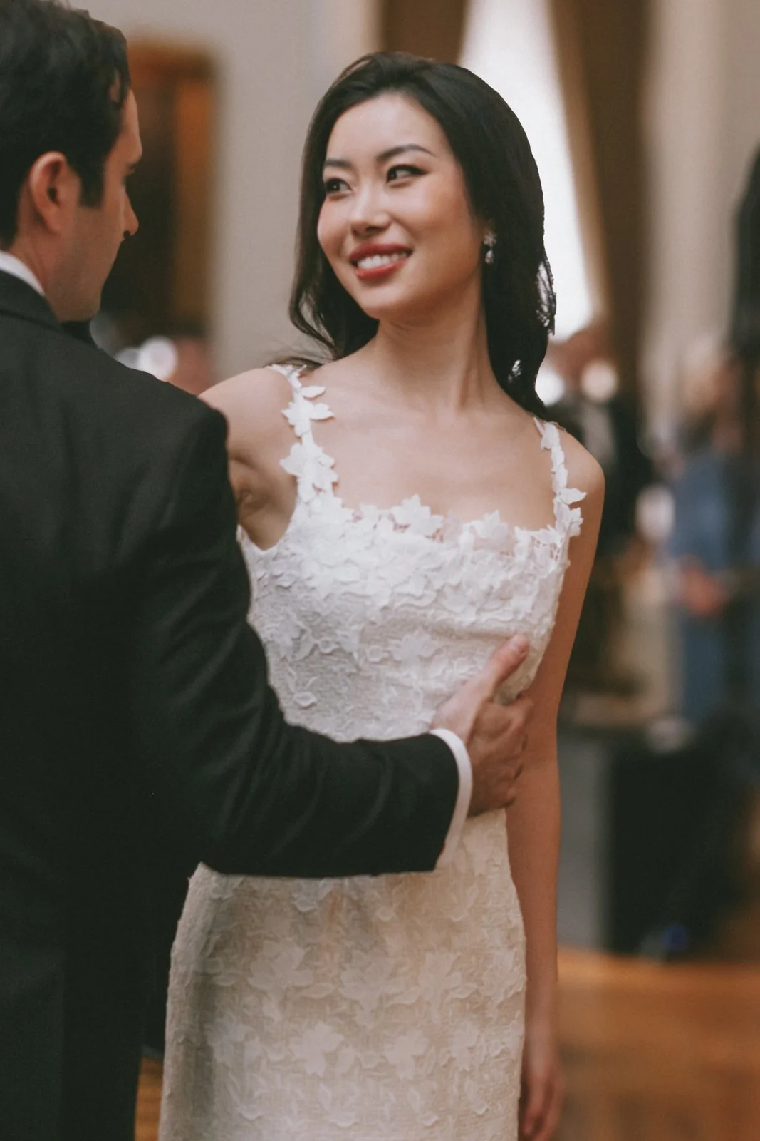A bride with dark hair and earrings, wearing a white lace wedding dress, smiling and looking at a man in a black suit who has his hand on her waist at a wedding ceremony.