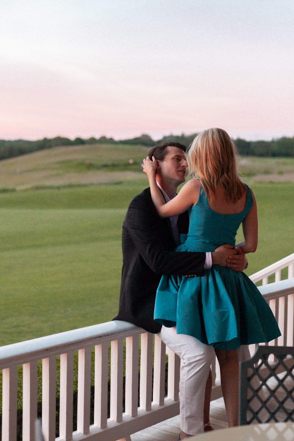 A man and a woman share an intimate moment on a porch, with the man sitting and the woman standing, surrounded by a scenic outdoor landscape at dusk.