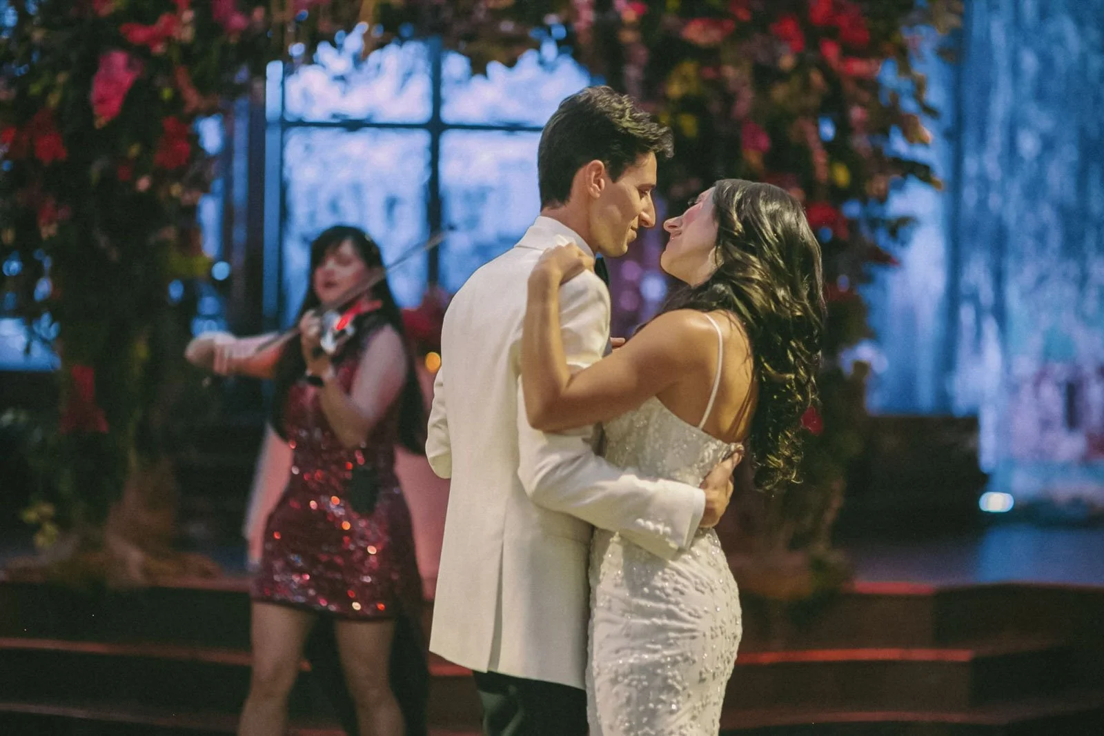 A couple in wedding attire dancing closely, a woman playing a violin in the background, decorated with red flowers, in a romantic indoor setting.