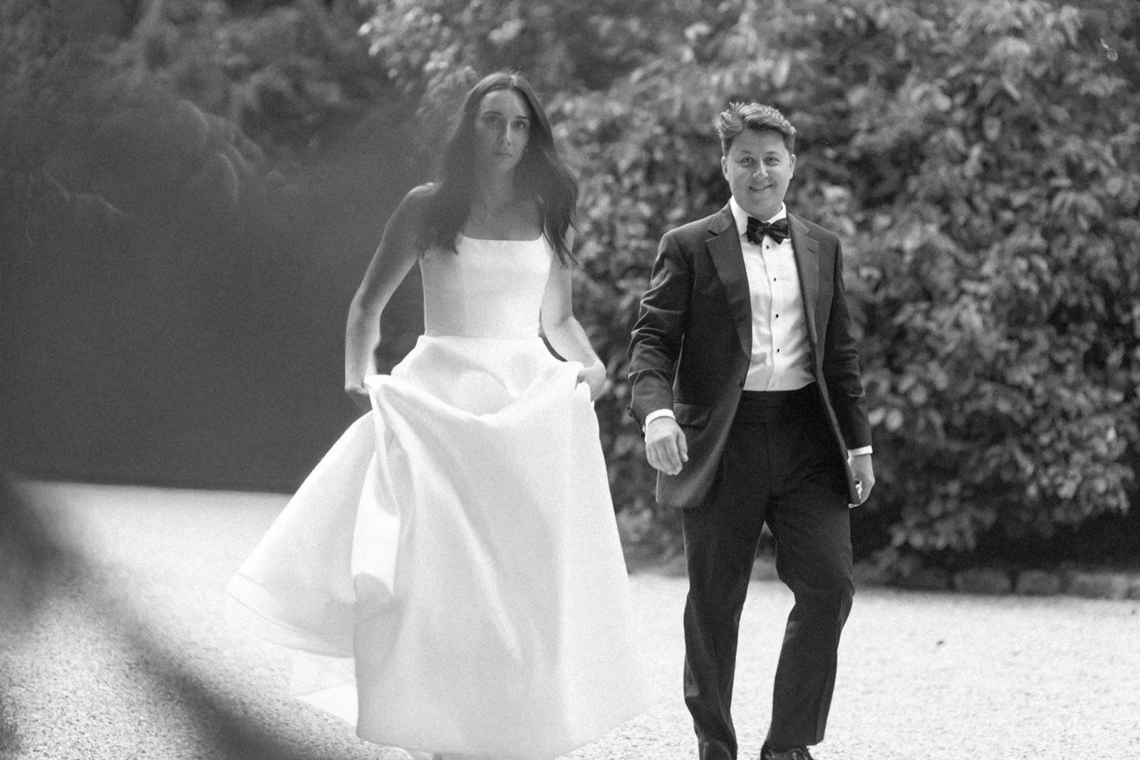 A black-and-white photo of a couple dressed in formal wedding attire walking outdoors, with the woman in a wedding dress and the man in a tuxedo with a bowtie, surrounded by trees.