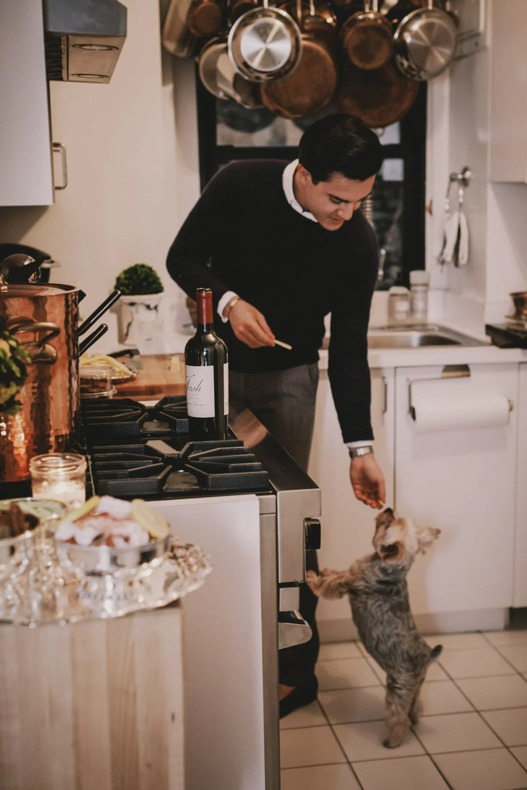 Man in a black sweater playing with a small dog in a kitchen, with hanging pots and pans on the wall.