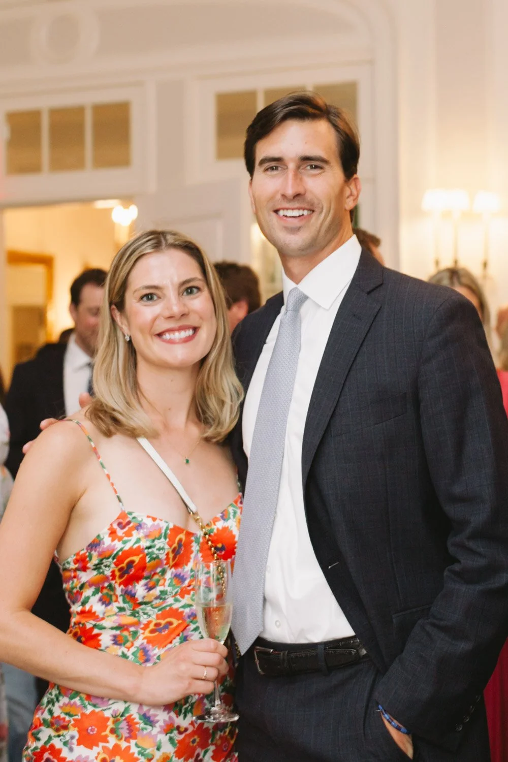 Two people, a woman in a floral dress holding a glass of champagne and a man in a suit at a social gathering or party.