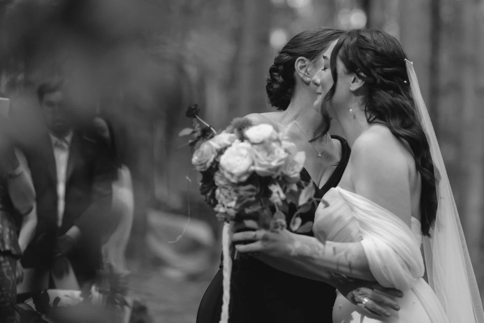 Two women, one with a bouquet, sharing a kiss at a wedding, with guests blurred in the background.