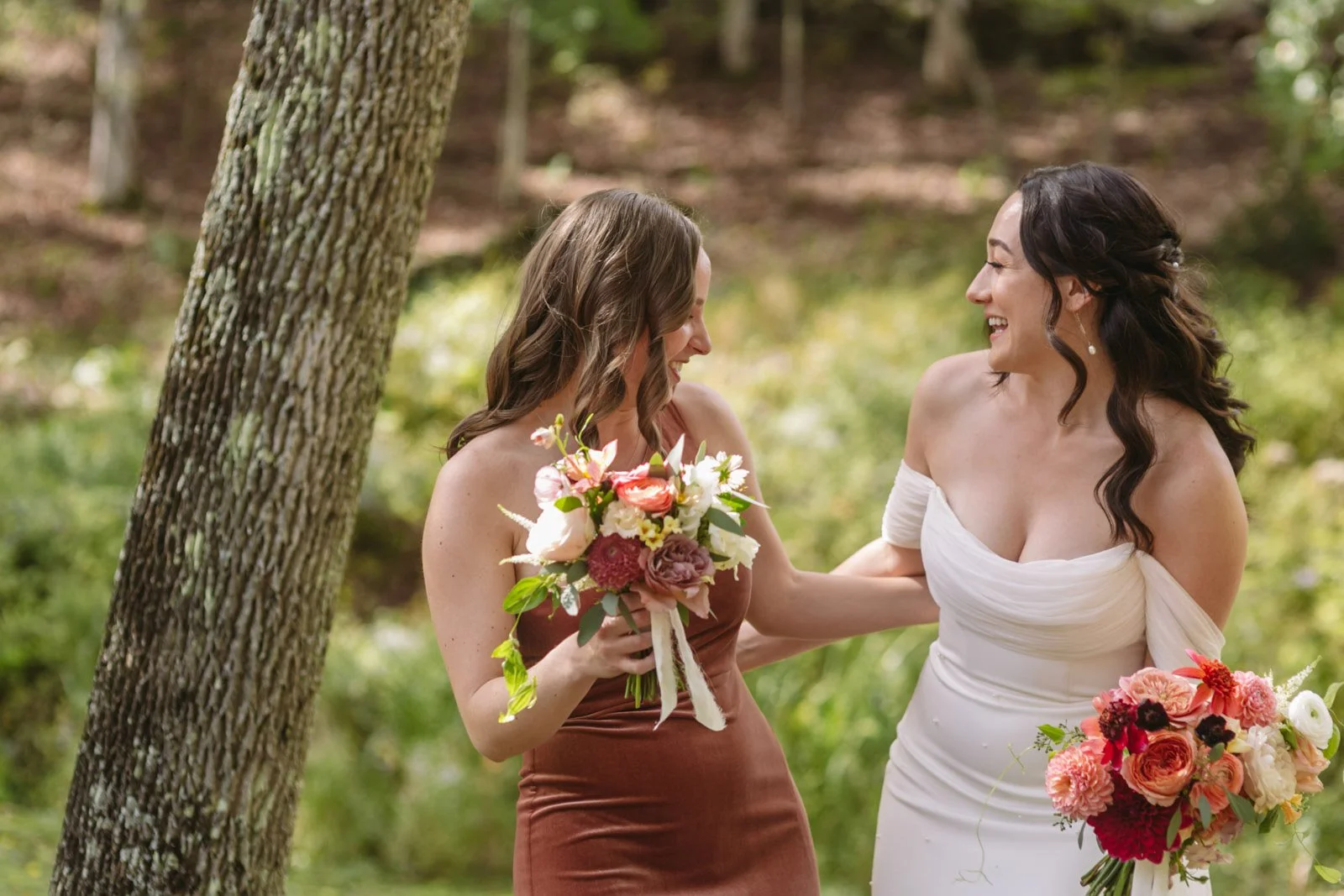 Two women standing outdoors, dressed in wedding attire, smiling and holding bouquets of flowers, with a tree in the foreground and greenery in the background.