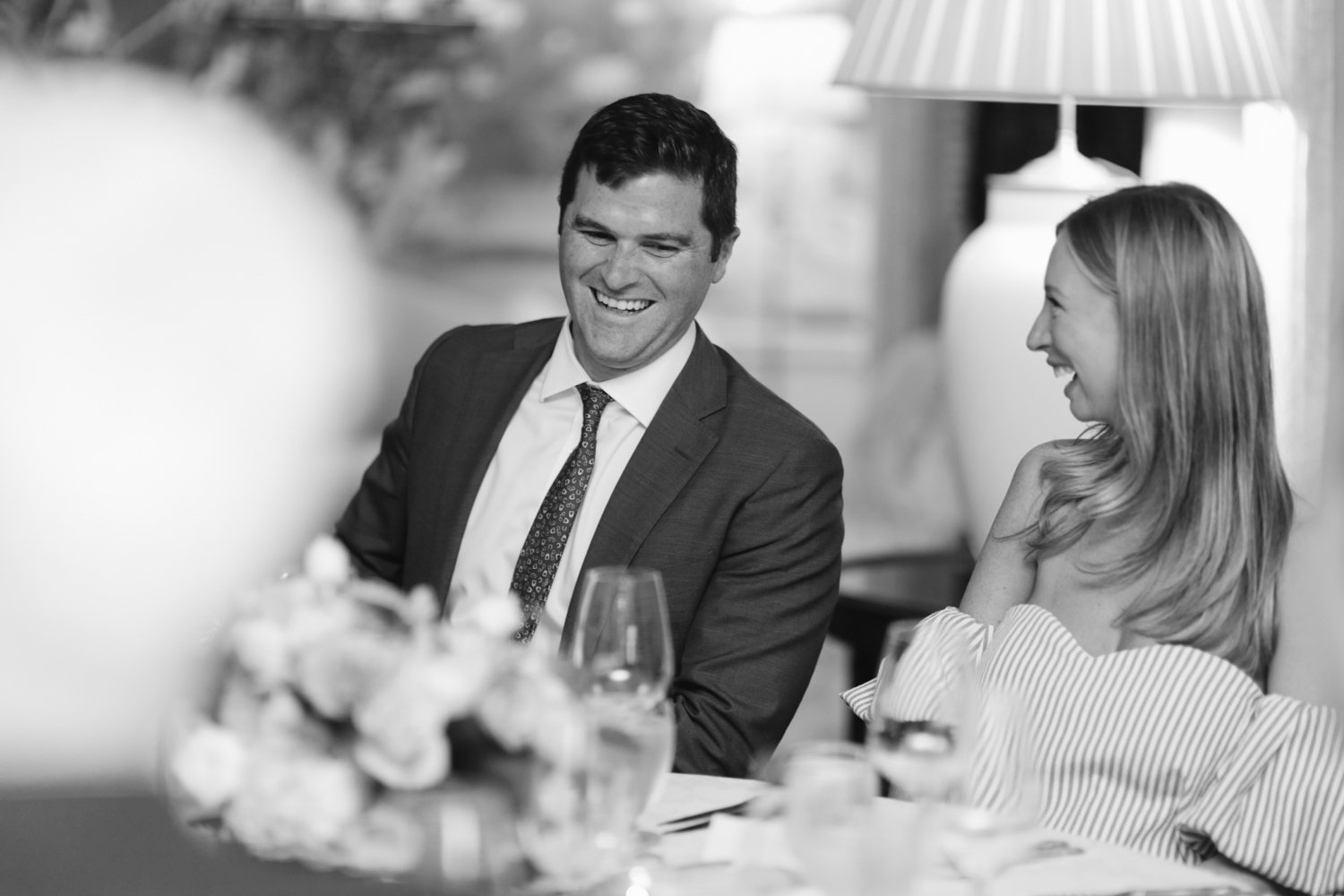 A black and white photo of a man and woman smiling and laughing at a social gathering, sitting at a table with floral centerpiece and wine glasses.