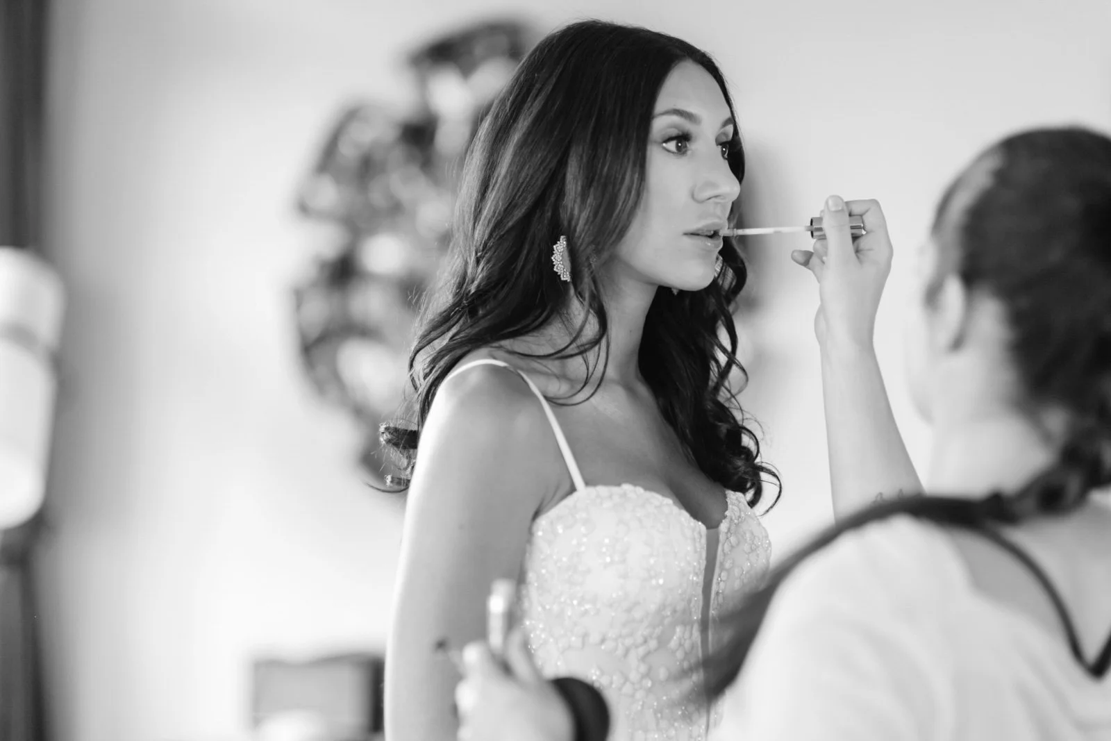 Black and white photo of a woman in a dress having makeup applied, with a makeup artist in the foreground.