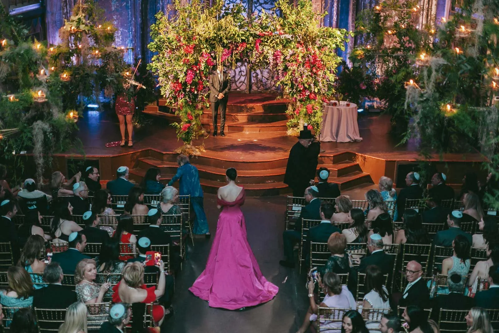 A wedding reception with a floral decorated stage, a woman in a pink gown walking towards the stage, guests seated in rows, and a man in a black hat and suit standing nearby.