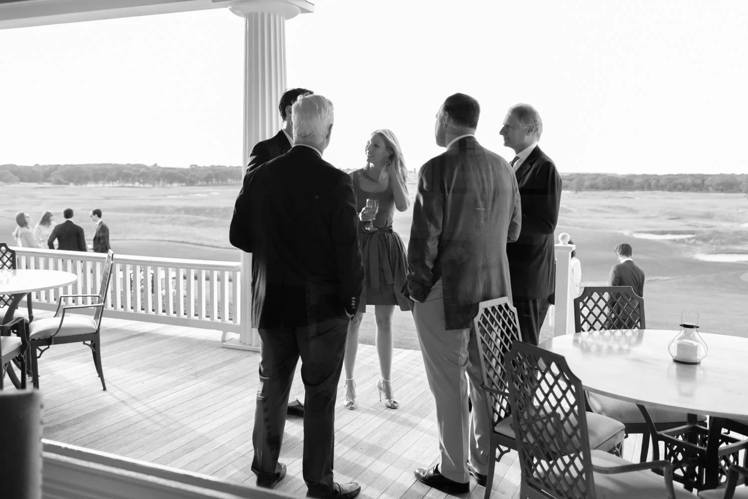 Black and white photo of a group of five people in formal attire standing on a porch, engaged in conversation with a scenic landscape in the background, including some people and a grassy field.