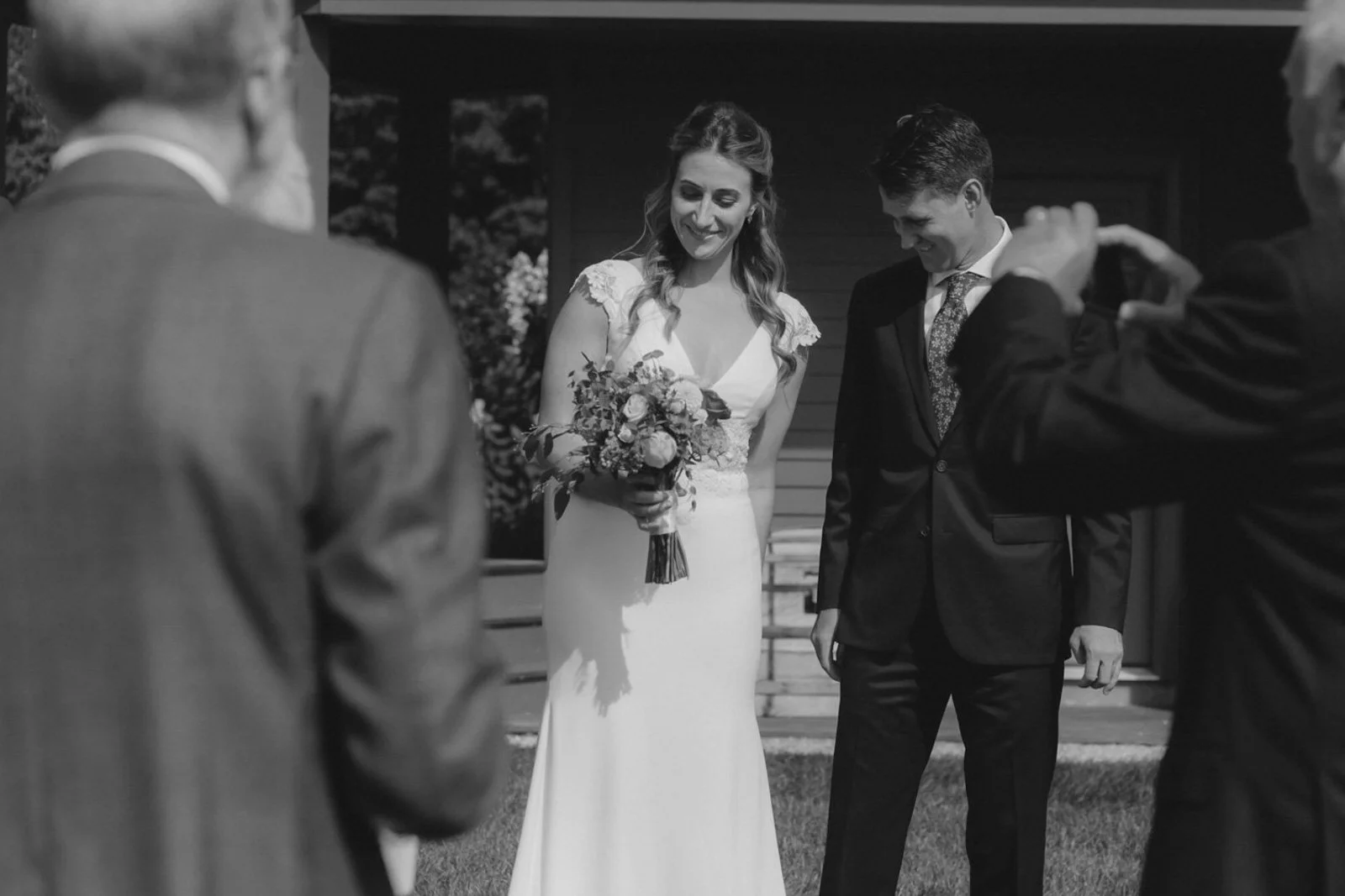 A woman in a wedding dress holding a bouquet, standing next to a man in a suit, during a wedding ceremony outdoors. Several guests are capturing the moment with cameras.