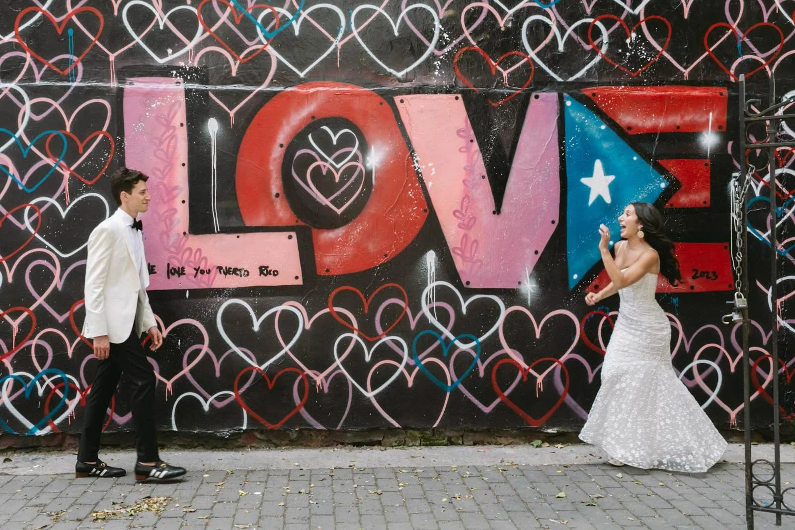 A man and a woman dressed in formal attire in front of a colorful graffiti wall with the word 'LOVE' and heart designs, smiling and facing each other.