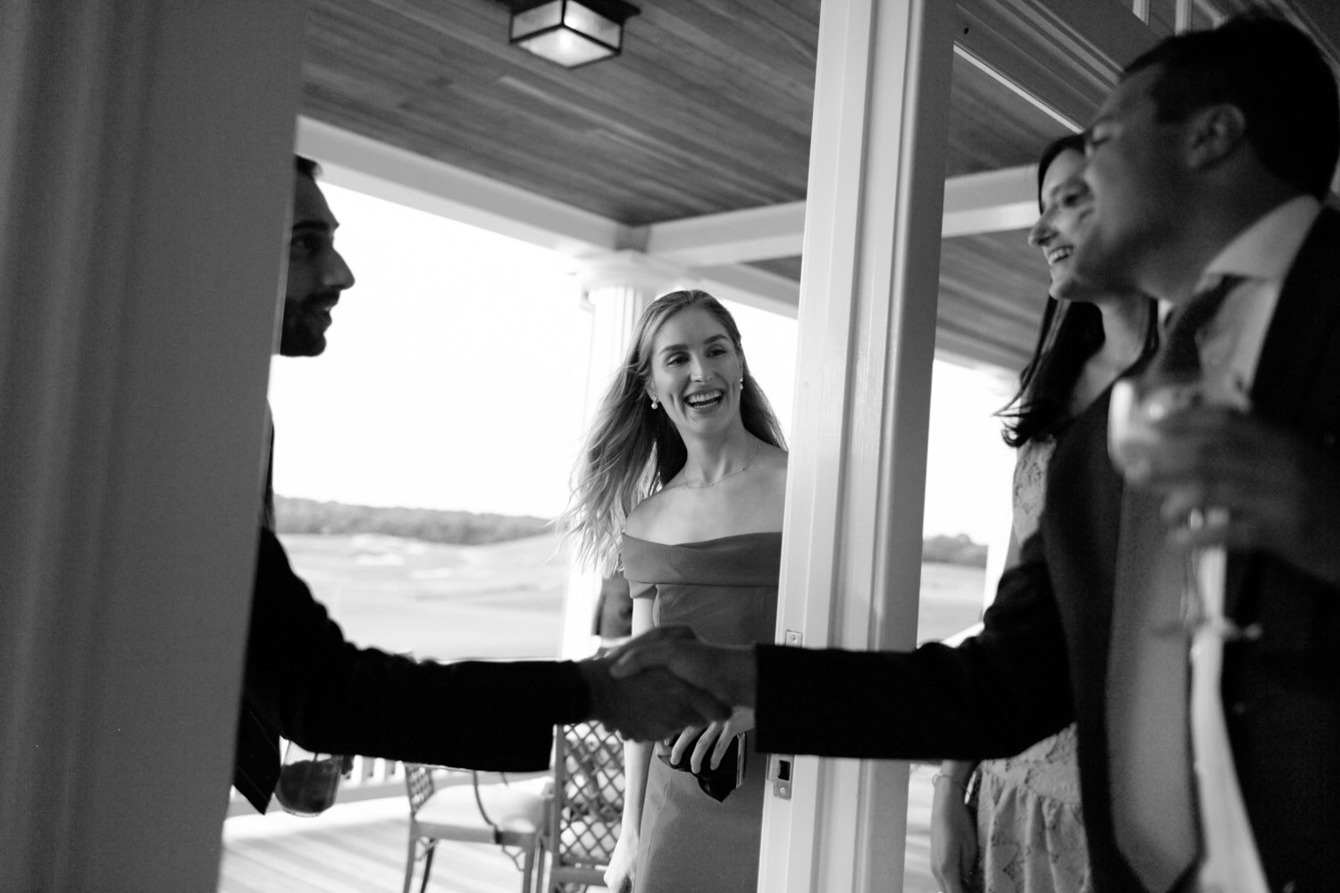 A woman in an off-shoulder dress smiling as she watches two men shaking hands at the doorway of a house, with a view of a deck and landscape in the background.