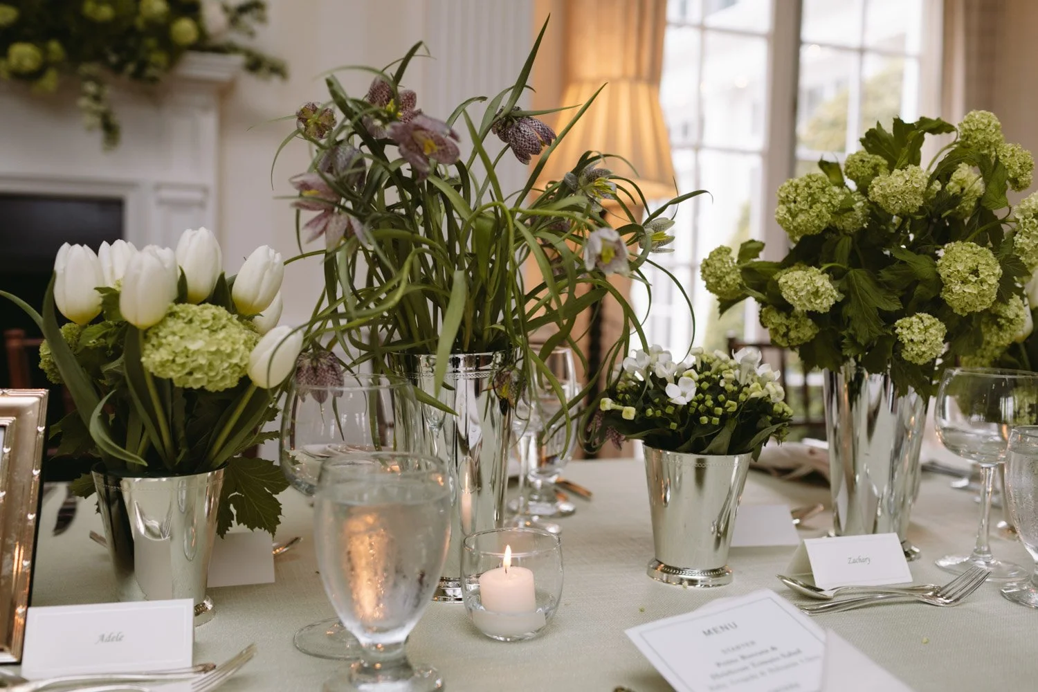 A table decorated with large floral arrangements in silver vases, smaller vases with white flowers, a lit candle in a glass holder, and place cards with names on a white tablecloth in a bright room with natural light.
