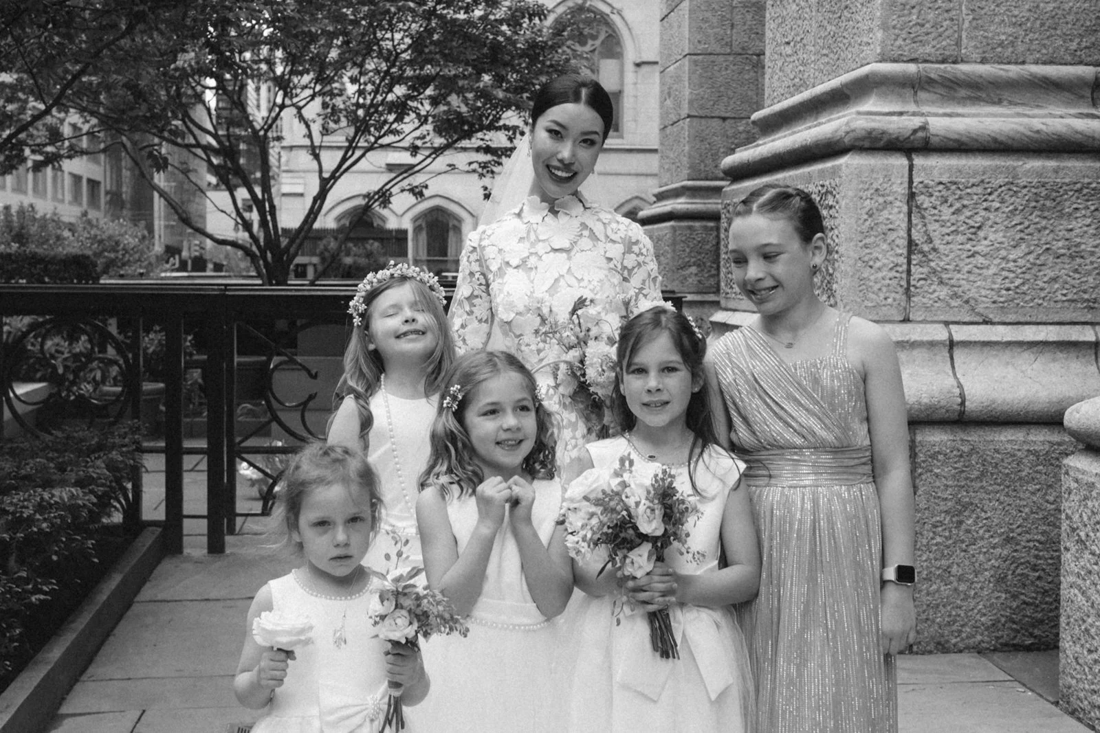 A group of six girls, including a woman, standing outside in front of a stone building and a tree, dressed in formal attire, with some holding flowers, smiling, in a celebratory setting.