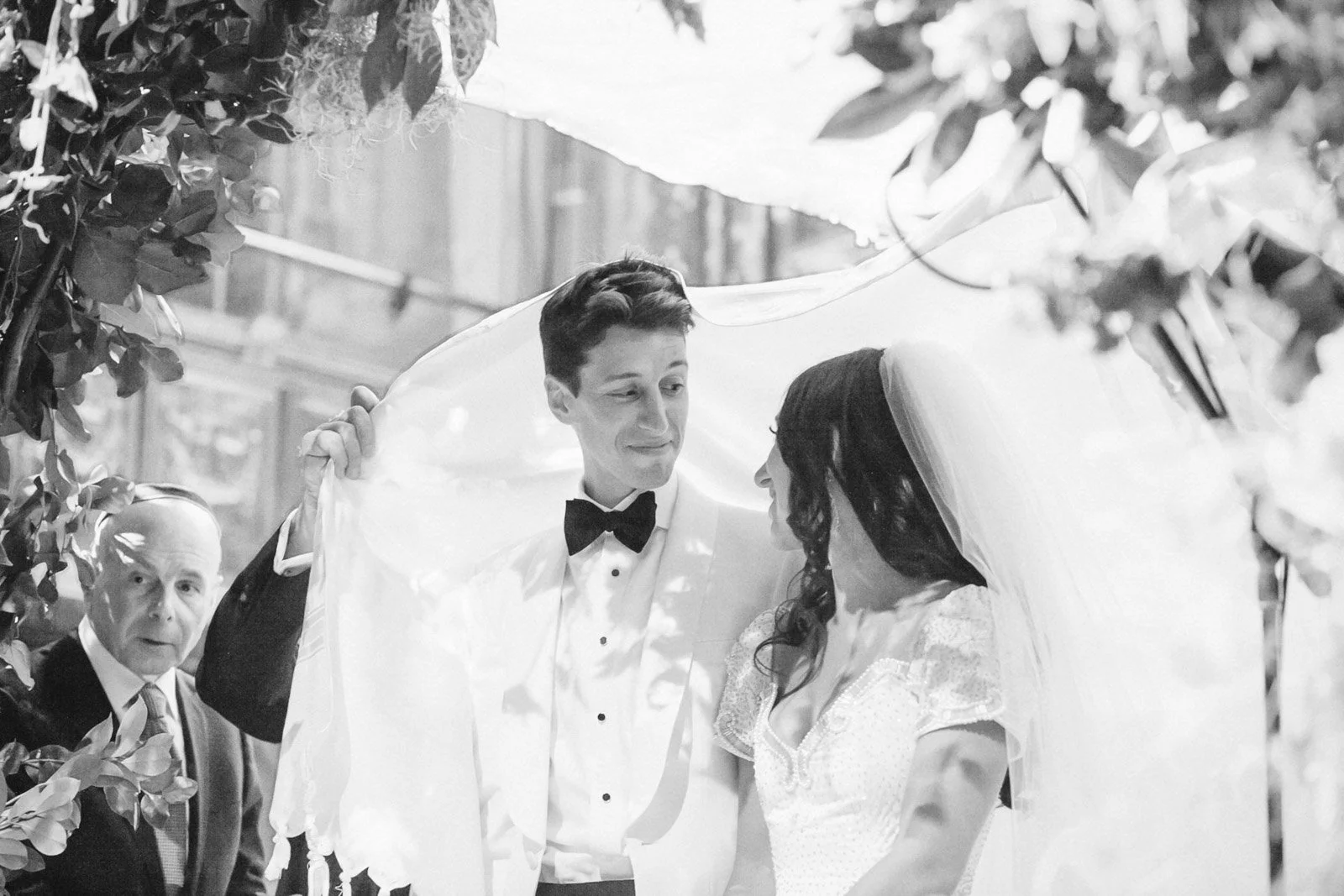A black and white photo of a wedding ceremony with a bride and groom, with the groom lifting the bride's veil, while a man looks on in the background.