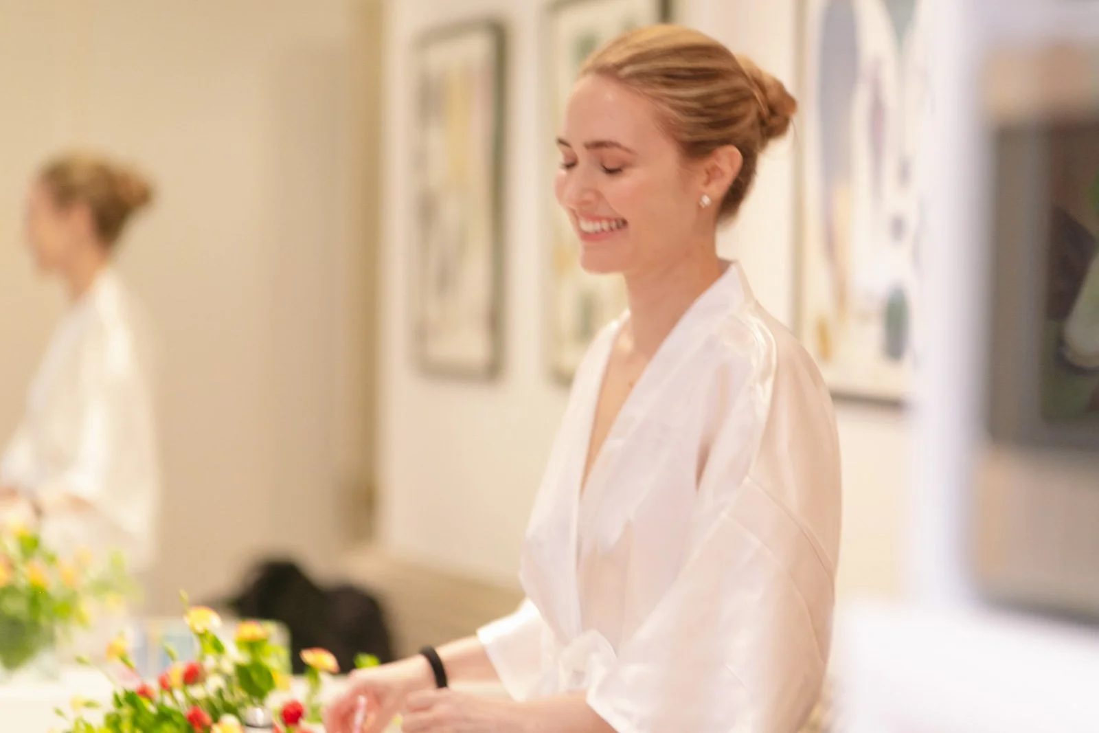 A bride smiling in a white silk robe, with her eyes closed, standing indoors with floral artwork on the wall behind her.