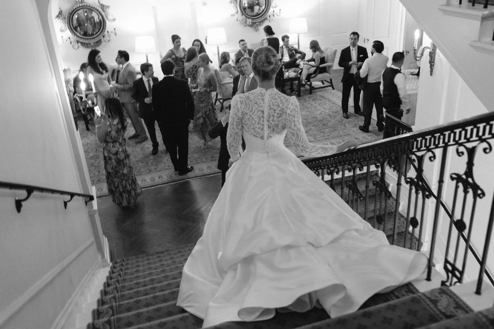 A bride in a white wedding gown with a lace top, standing on a staircase overlooking a social gathering in a decorated elegant room at The Colony Club New York.