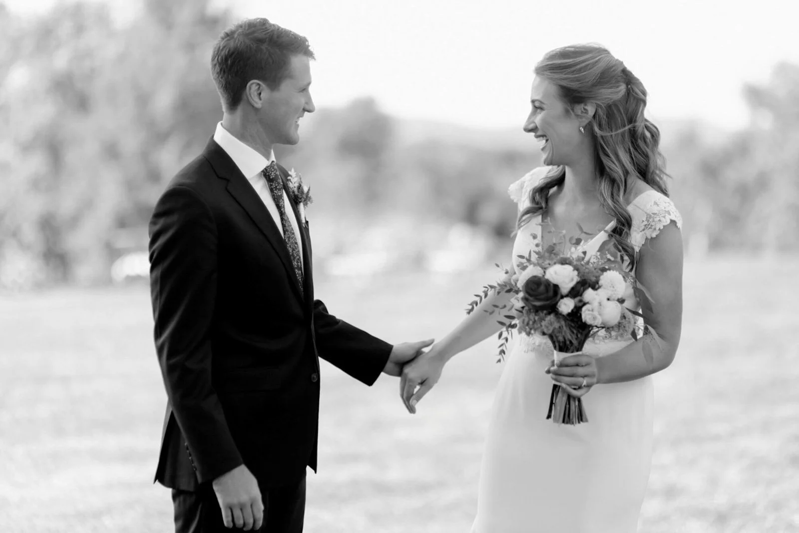 Black and white photo of a bride and groom holding hands and smiling at each other outdoors.