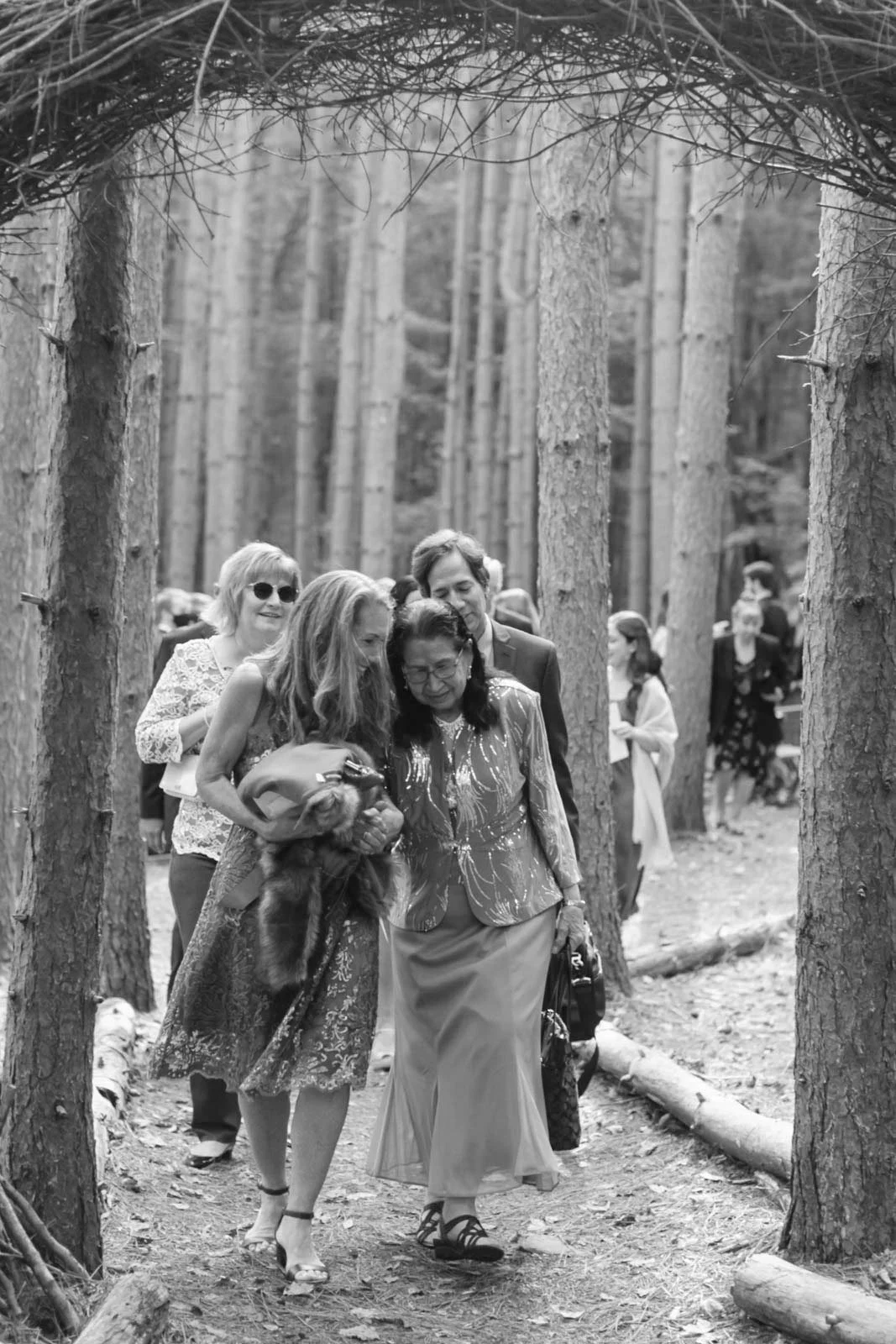 Group of people walking through a wooded forest trail. The woman in front is dressed in elegant clothing and holding a fur stole, while the woman beside her is wearing a patterned blazer and skirt. Other people are visible in the background among tal