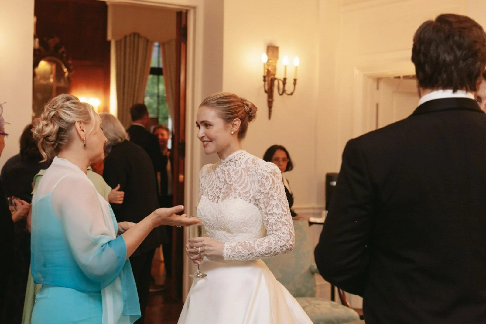 A bride in a white lace wedding dress and updo holds a glass of champagne, smiling and appears to be engaged in conversation with an older woman in a colorful dress at a wedding reception.
