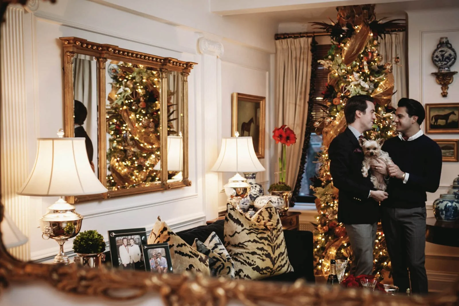 Two men in formal attire standing in a decorated living room near a Christmas tree, holding a small dog, with festive decorations and family photos on a patterned cushion sofa.