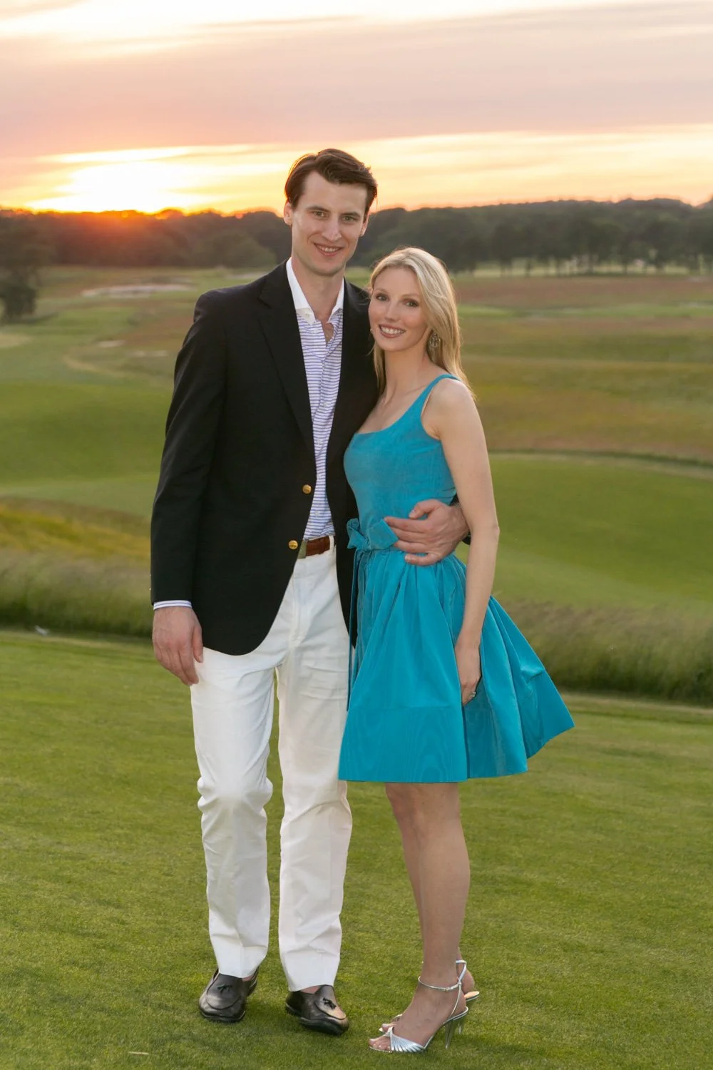 A young man and woman stand together on a golf course during sunset, dressed in semi-formal attire, smiling at the camera.