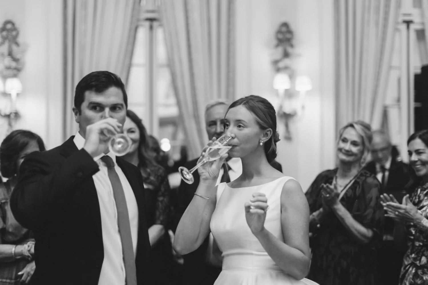 Black and white photo of a wedding reception with a woman in a white dress and a man in a suit drinking champagne while surrounded by smiling guests in a decorated elegant room.