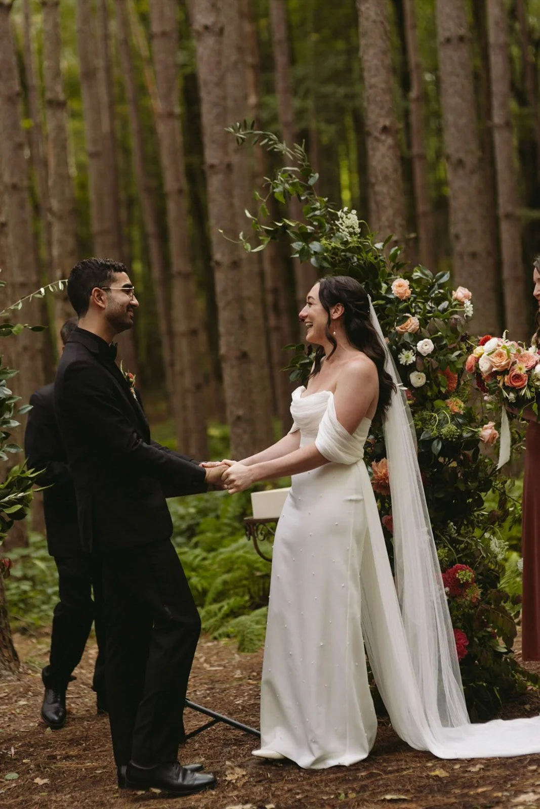 A couple getting married outdoors in a forest, holding hands, with a floral arch behind them.