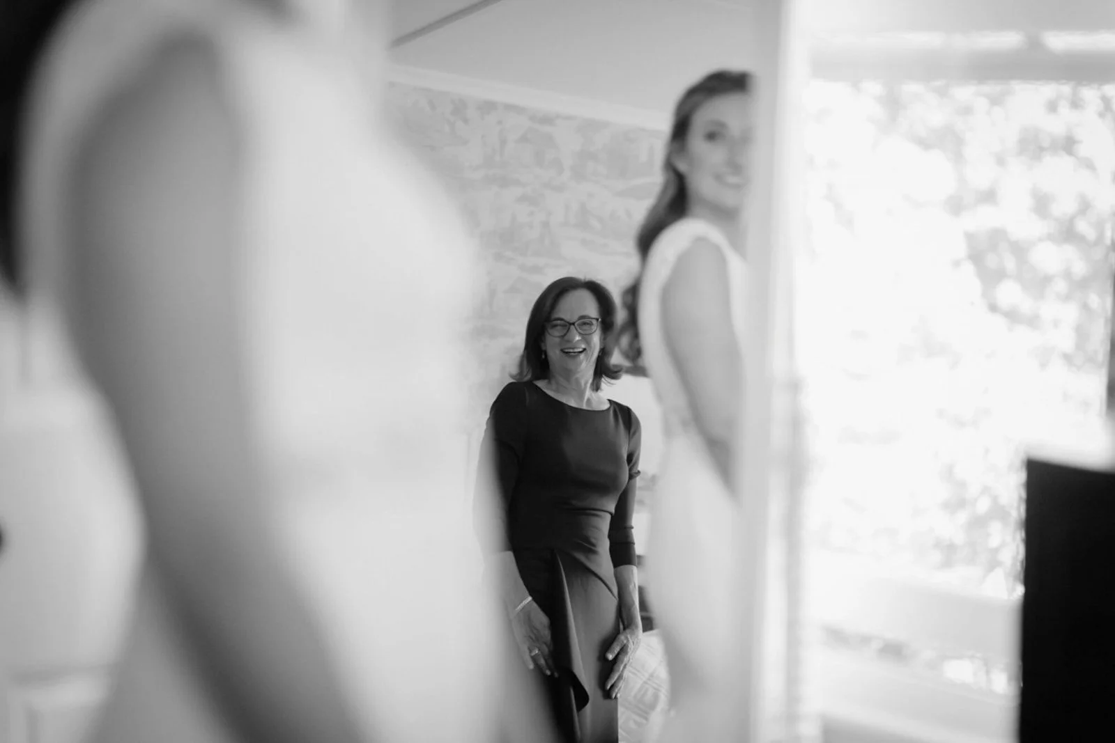 A woman smiling and standing in front of a window, seen through a mirror with out-of-focus foreground elements, in black and white.