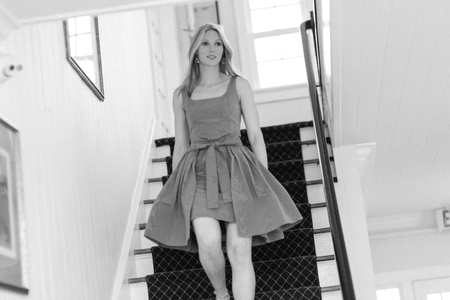 A woman in a sleeveless dress with a tied waist walks down a staircase, smiling and looking ahead. The staircase has a patterned carpet runner and is inside the Shinnecock Golf Club.