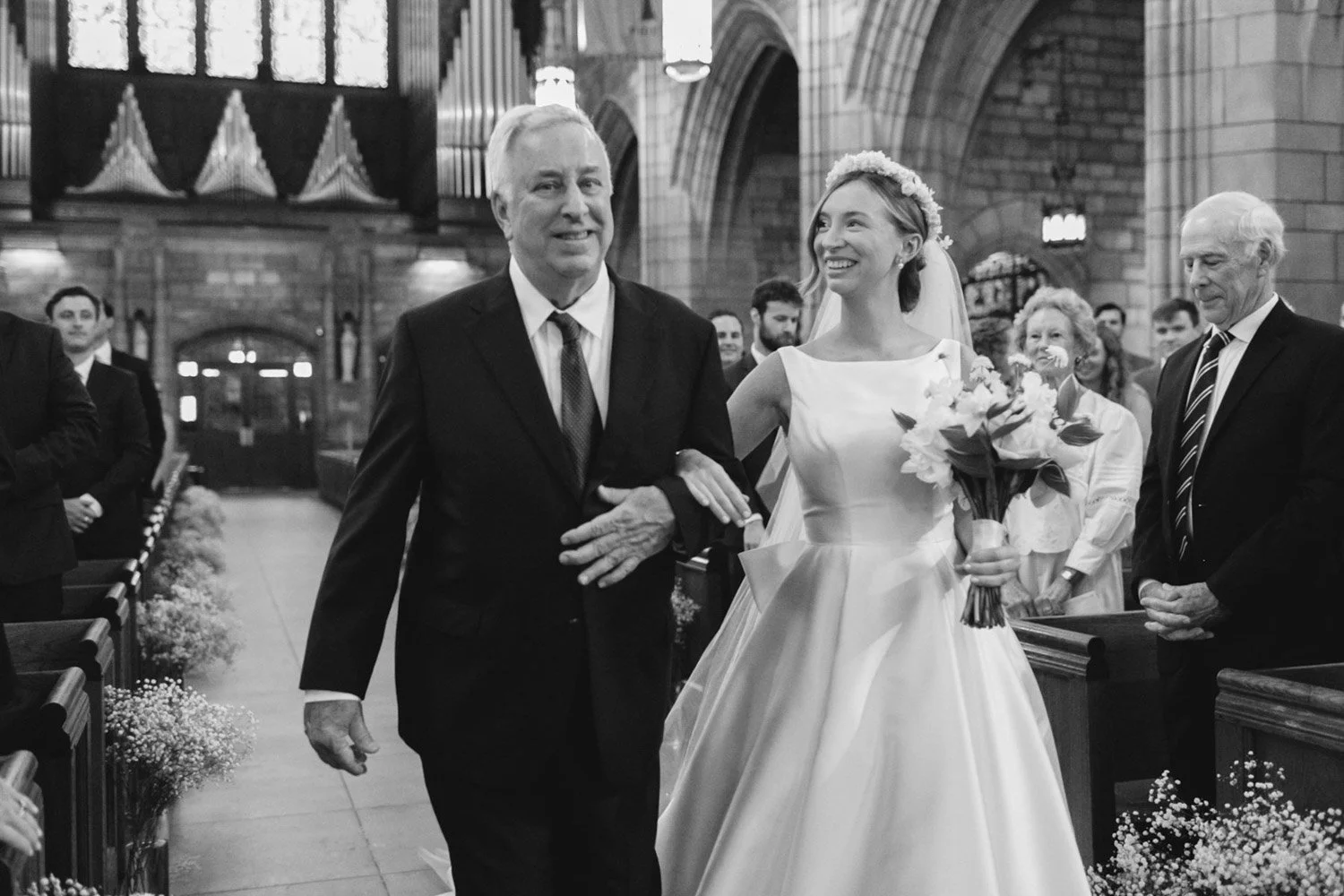 A bride in a wedding dress holding a bouquet of flowers walking down the aisle with an older man, likely her father, inside a church. Other guests are visible in the background.