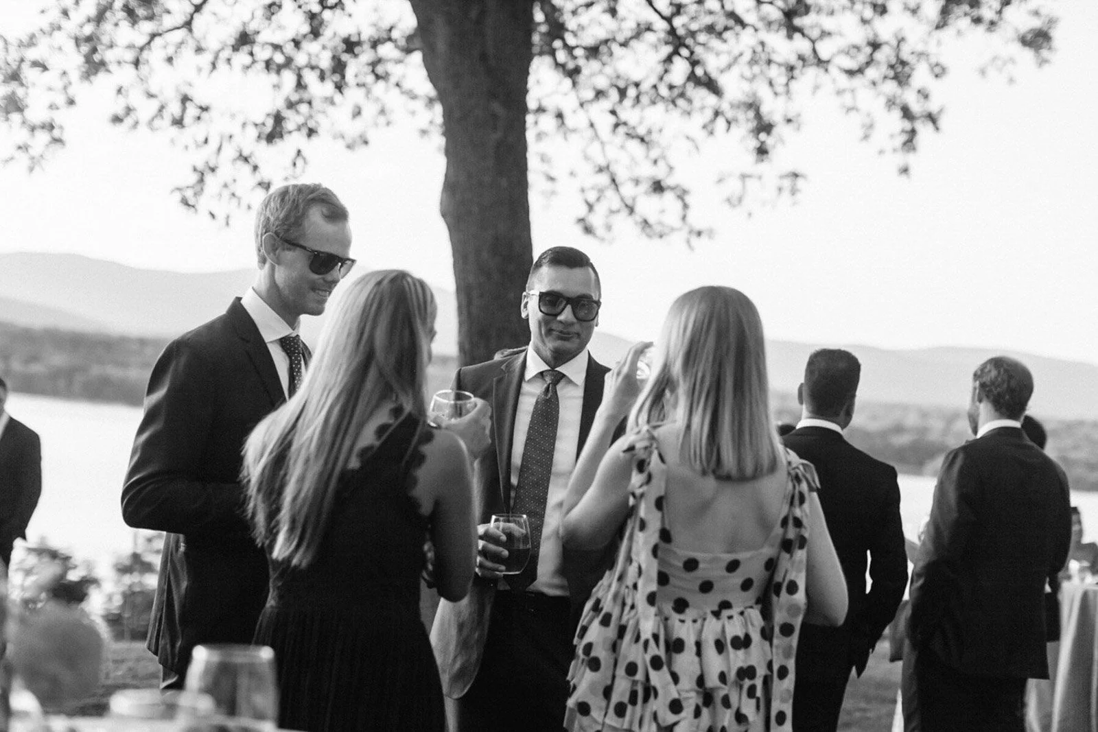 People socializing outdoors near a lake or river with trees and hills in the background. They are wearing formal or semi-formal attire and holding drinks, suggesting a social event or gathering.