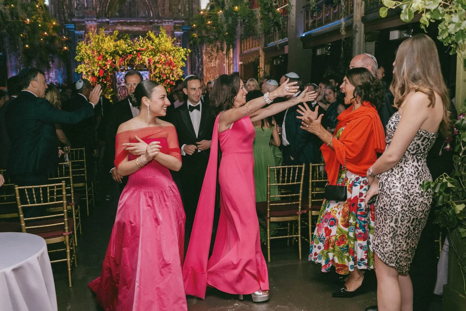 A group of people at a celebration, with women dressed in colorful dresses, including pink and animal print, interacting and smiling in a lively, decorated venue with flowers and string lights.