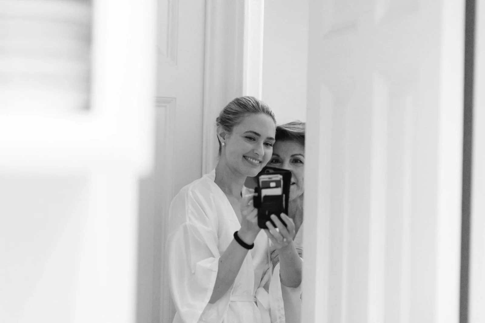Two women taking a selfie in front of a mirror, smiling. One woman is holding a smartphone, capturing the photo.