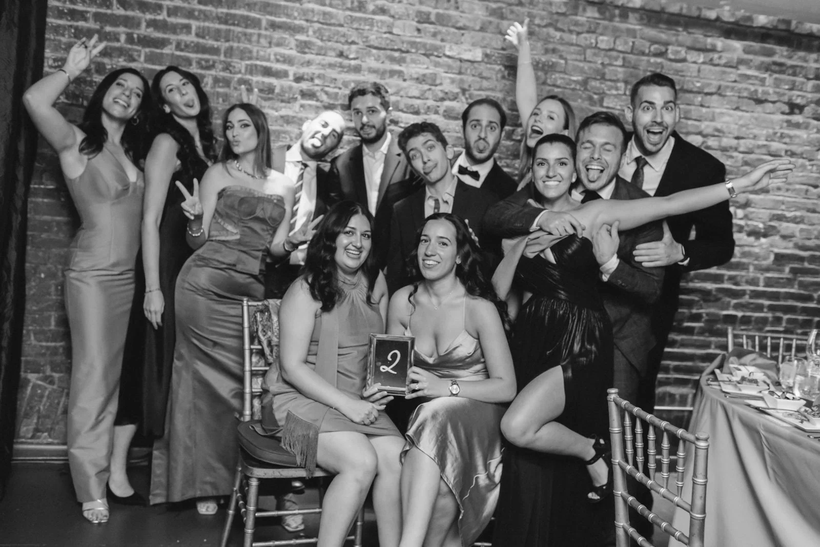 A group of people dressed in formal attire at a celebration, with some sitting and others standing, laughing and making silly faces in front of a brick wall.