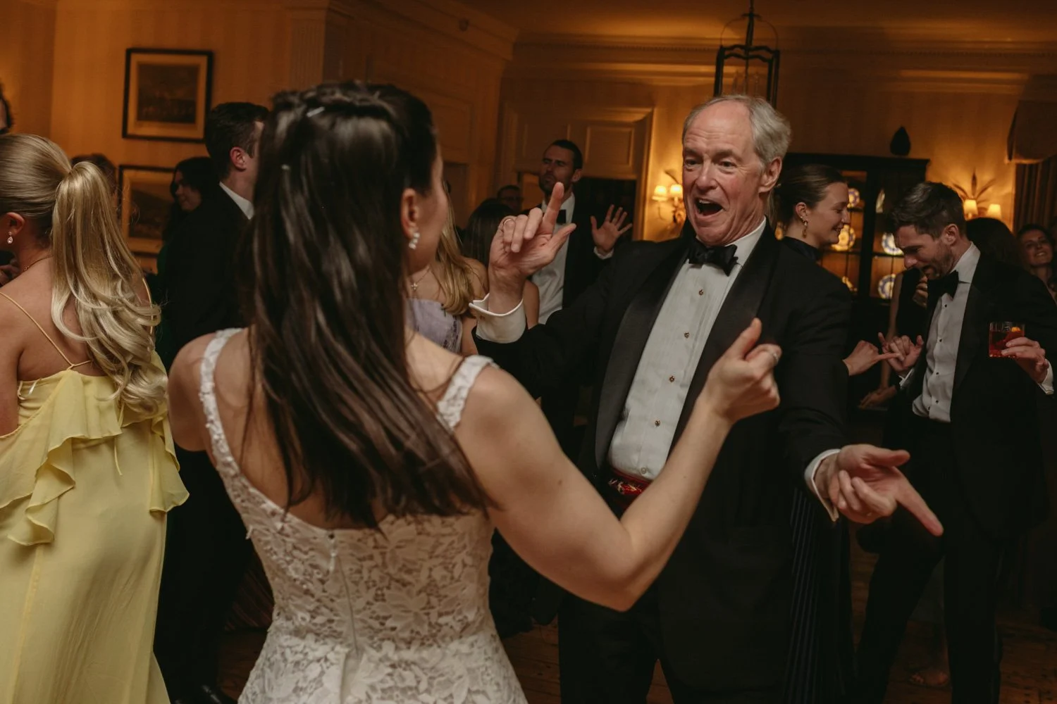 Older man and young woman dancing at a formal event, surrounded by other people dancing and socializing in an elegant room.
