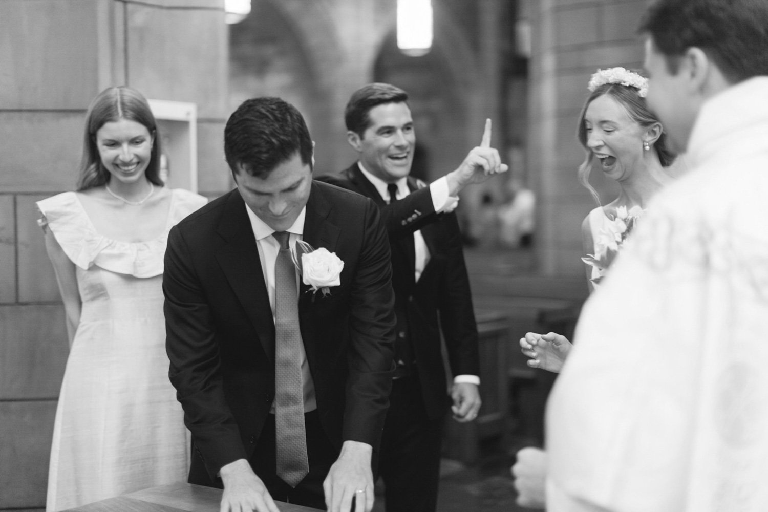 Black and white photo of a wedding ceremony capturing a man tying a ring on a woman's finger, surrounded by joyful friends and family.