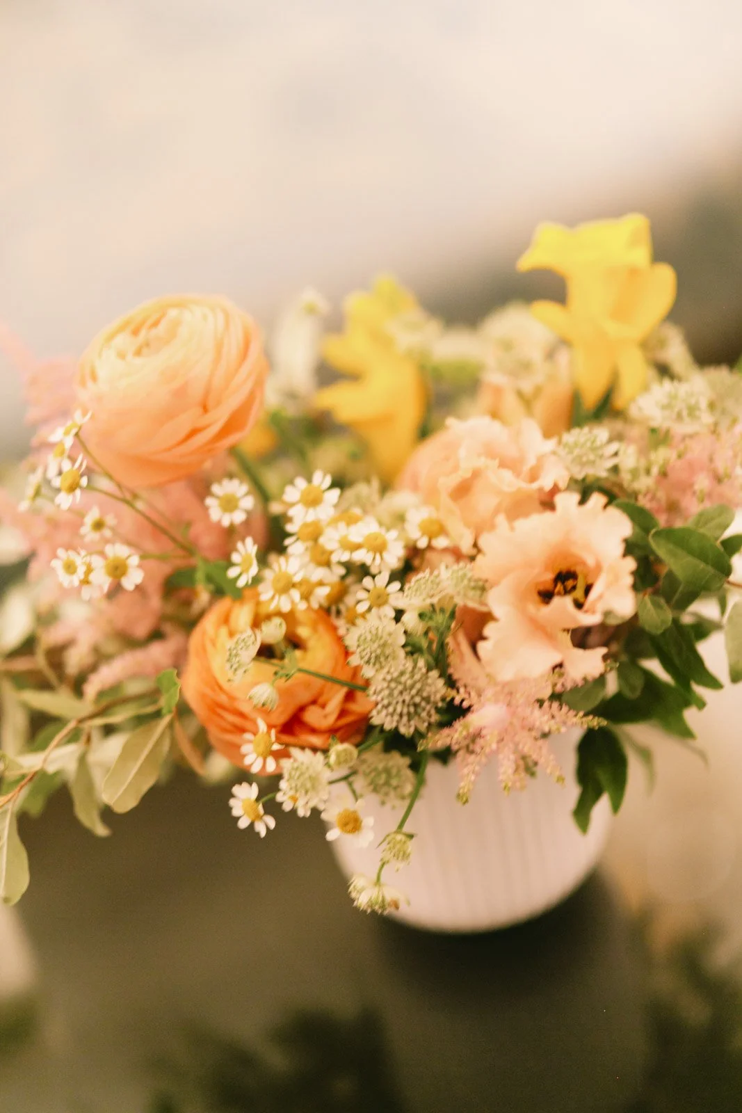 A bouquet of mixed flowers including orange ranunculus, pink lisianthus, yellow daffodils, and small white daisies in a white vase.