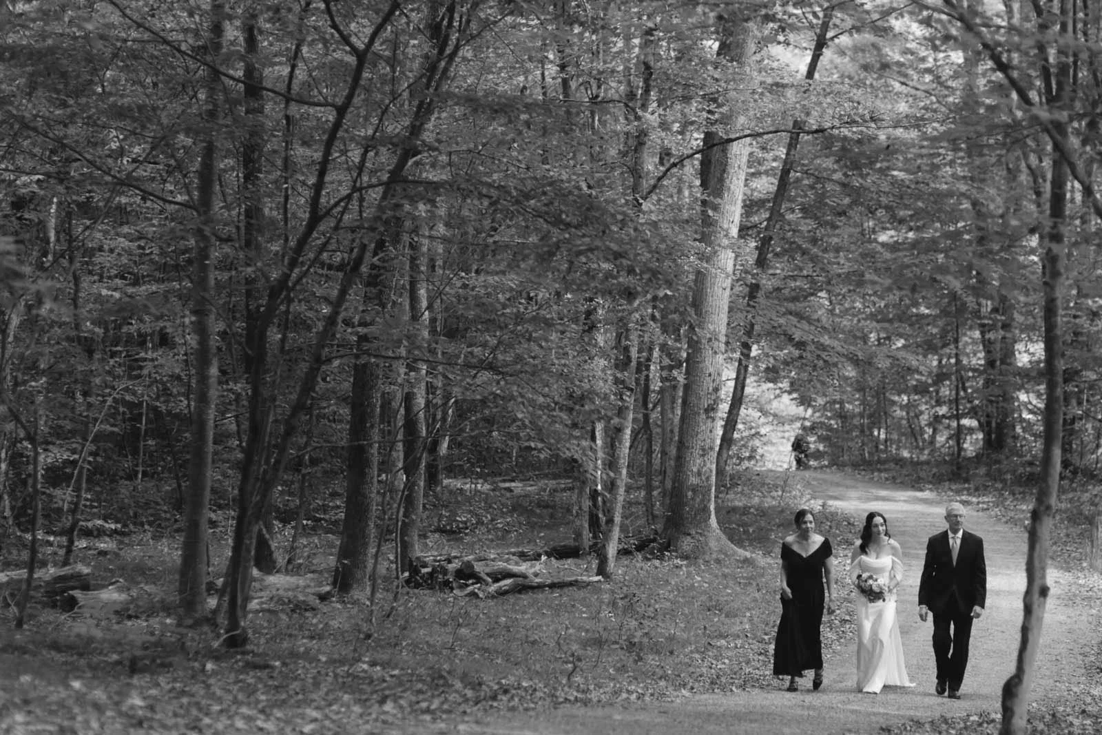 A black and white photo of a wedding party walking on a dirt path through a wooded forest, with a bride in a white gown holding a bouquet, a groom in a suit, and a woman in a dark dress.