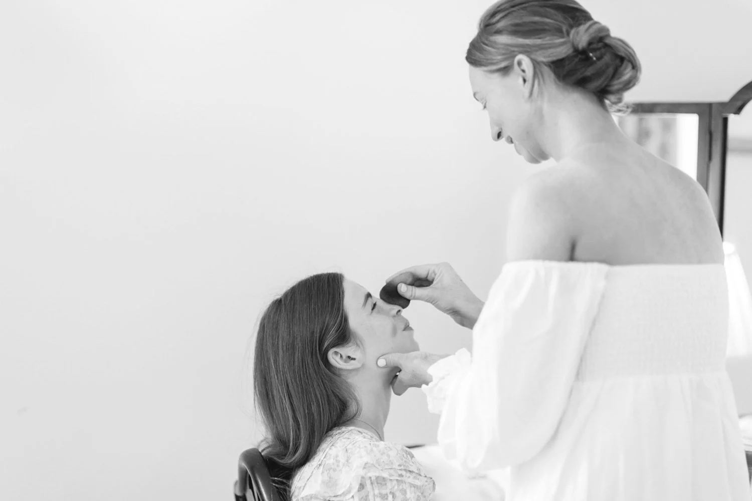 A woman in a white gown is applying makeup on a seated woman with long dark hair, in a room with a plain wall.