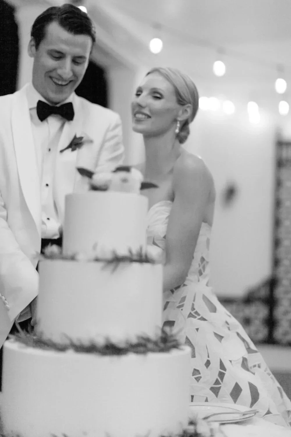 A black-and-white photo of a bride and groom cutting a wedding cake together, smiling and celebrating at their wedding reception.