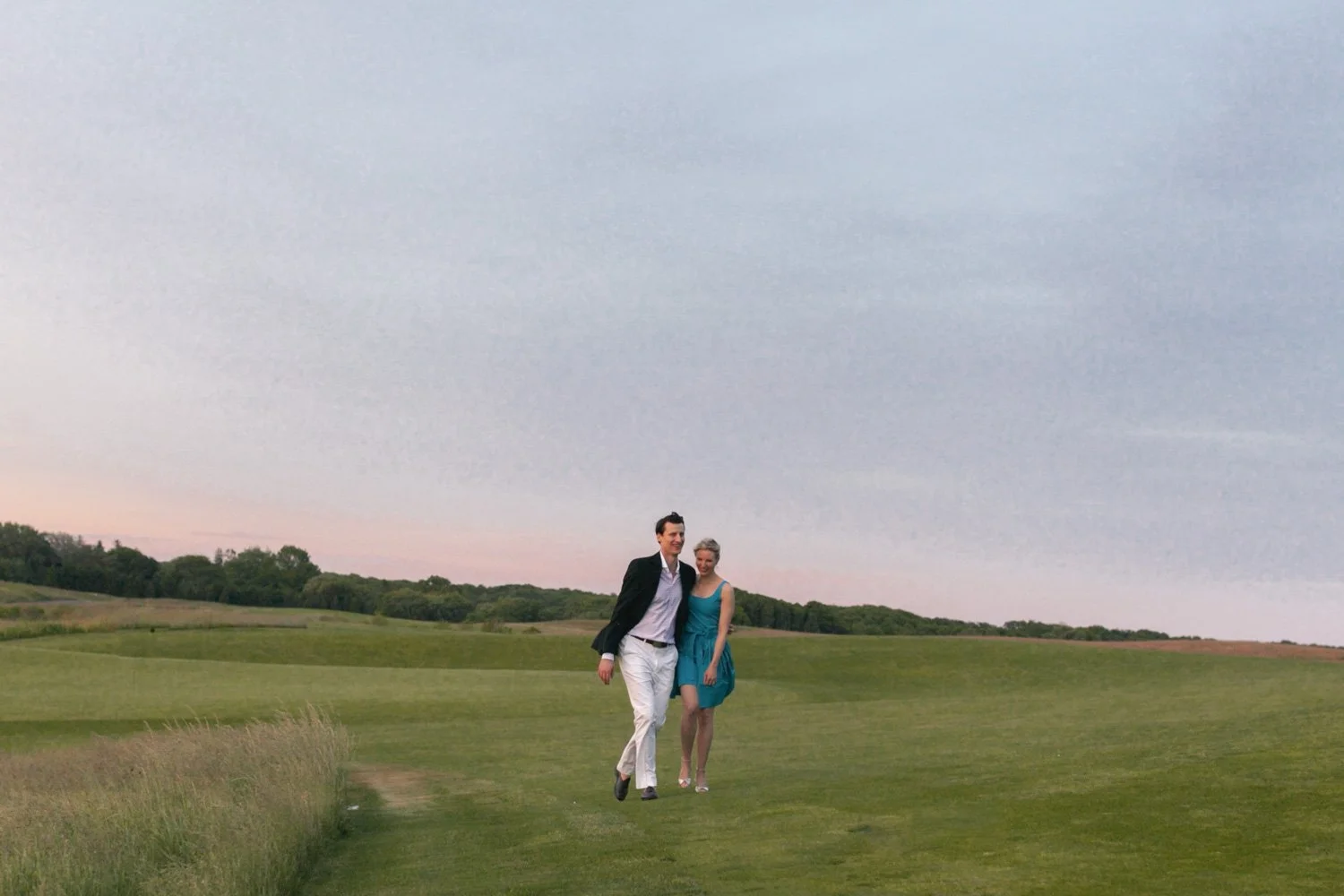 A man and woman walking together outdoors on a grassy field during dusk, with a landscape of rolling hills and a cloudy sky in the background.