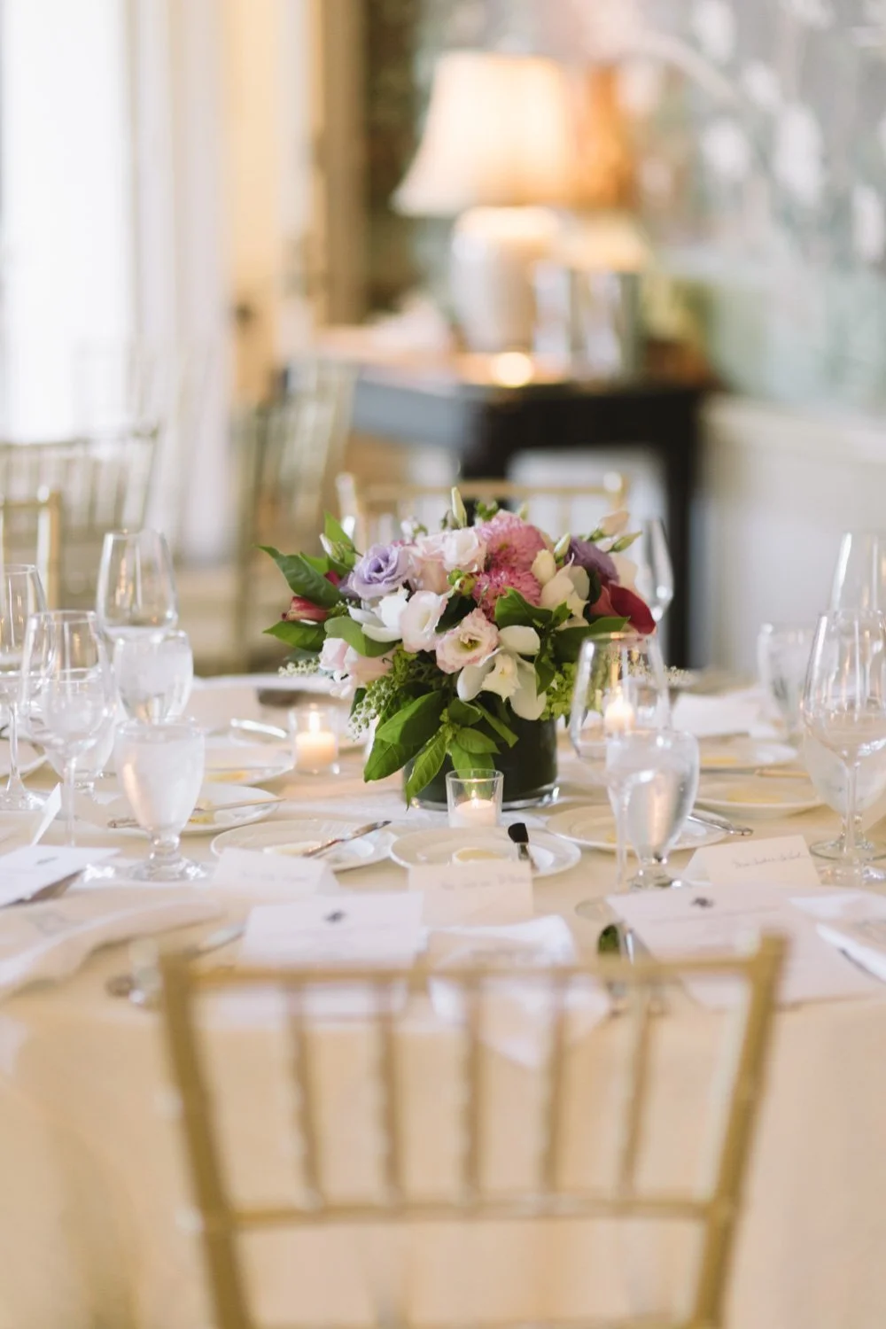 Elegant table setting with a floral centerpiece of pink, white, and purple flowers, surrounded by water glasses, candles, and place cards in a well-lit room.