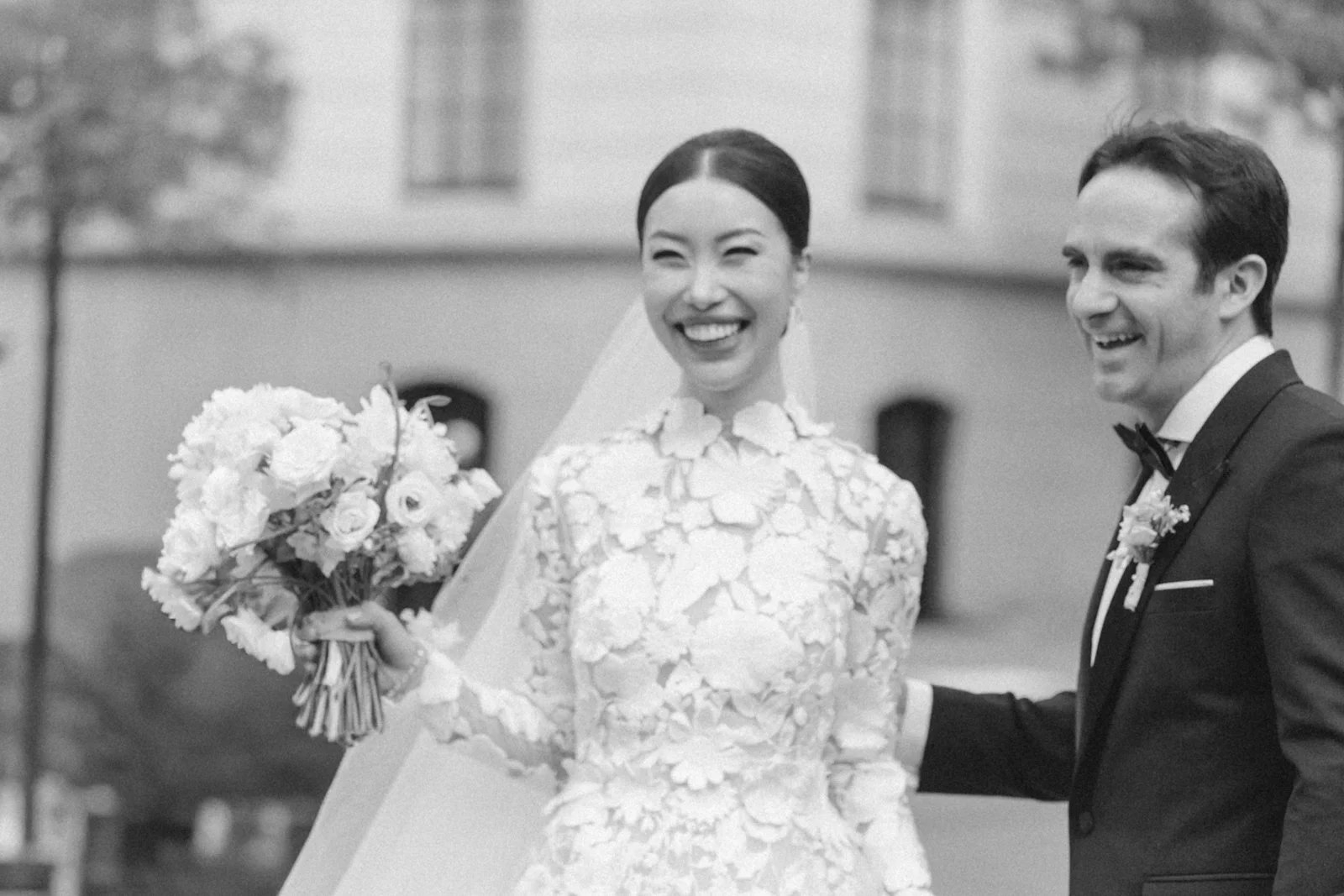 Black and white photo of a smiling bride in a lace wedding gown holding a bouquet of flowers, and a groom in a tuxedo with a bow tie, smiling and holding her hand outdoors, with trees and buildings in the background.