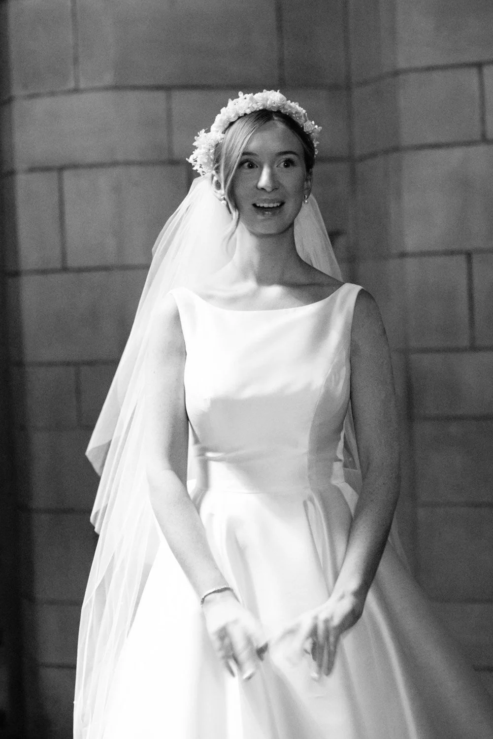 A black and white photo of a woman dressed in a wedding gown with a veil, wearing a floral headband, standing indoors against a brick or stone wall background.