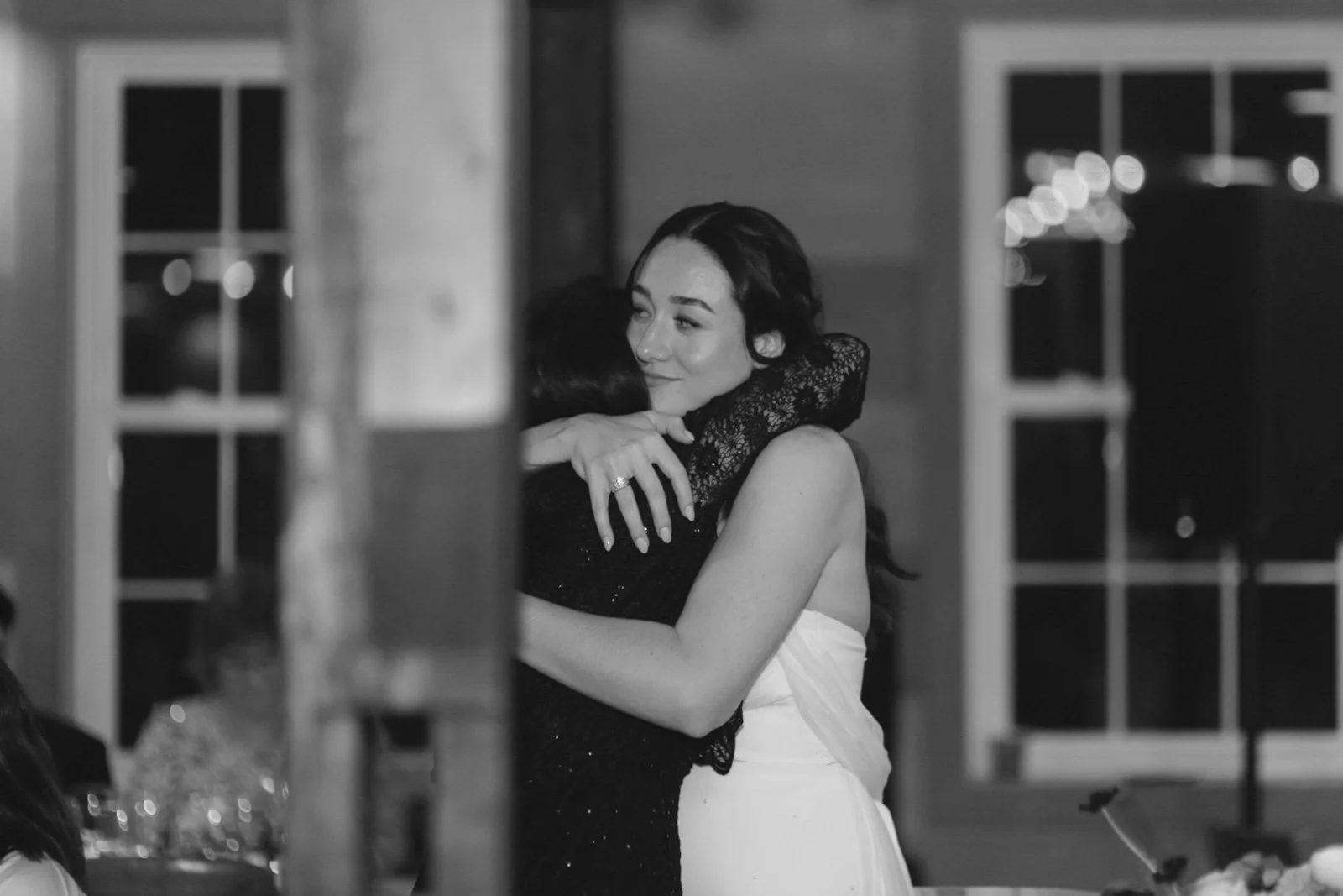 Two women hugging, one in a white dress and the other in a black lace dress, at a social gathering or wedding reception in black and white.