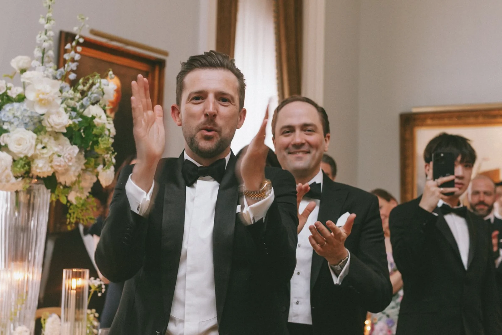 Two men in tuxedos at a formal event, one with hands raised, surrounded by others, including a person taking a photo with a smartphone, in a decorated room with flowers and candles.