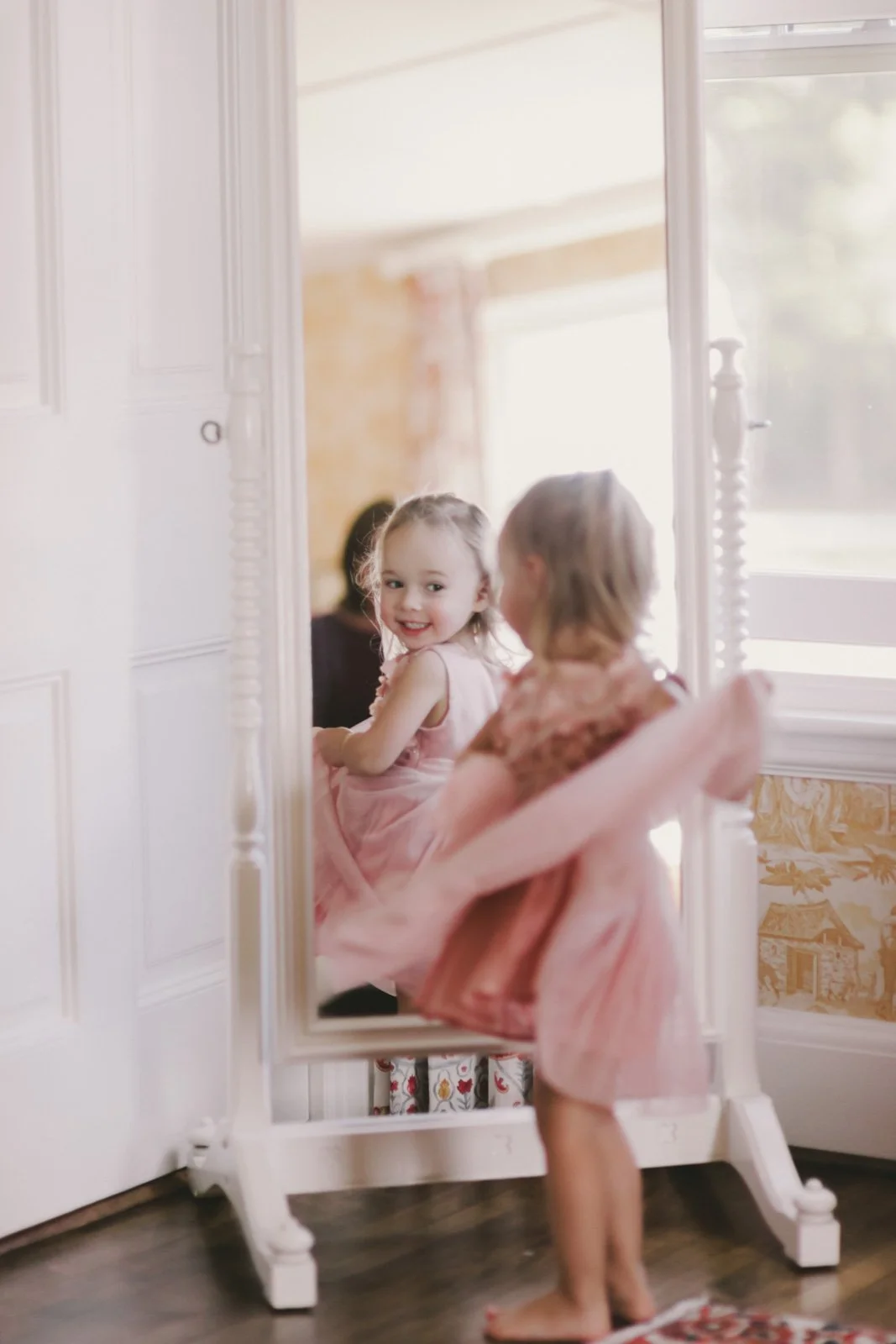 A young girl in a pink dress sits on a chair and looks into a large mirror, smiling at her reflection. The mirror rests on a white stand, with sunlight coming through a nearby window illuminating the scene.