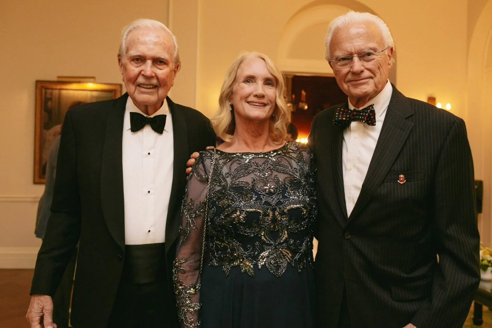Three elderly people in formal attire standing close together in a warmly lit room, smiling for the camera.